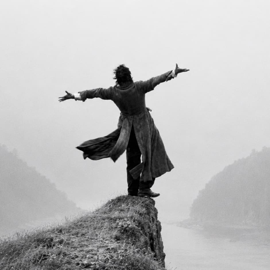 A lone figure standing with arms outstretched on a clifftop above a misty river valley, in black and white