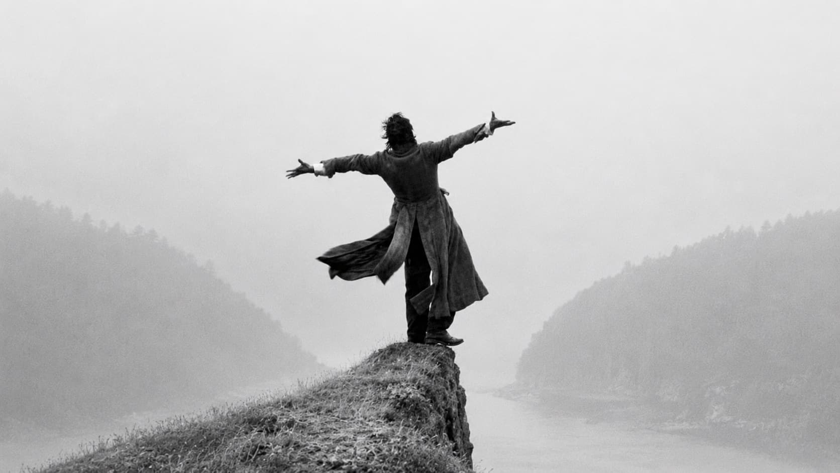 A lone figure standing with arms outstretched on a clifftop above a misty river valley, in black and white