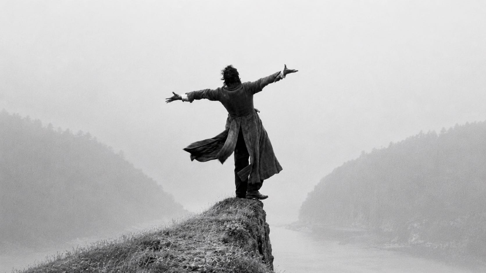 A lone figure standing with arms outstretched on a clifftop above a misty river valley, in black and white